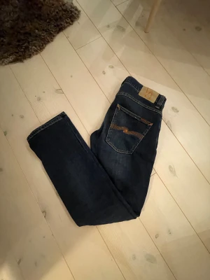 Mörkblå jeans från Nudie Jeans Co W30 L32 - Snygga mörkblå jeans från Nudie Jeans Co med klassisk femficksdesign och orangea sömmar på bakfickorna. Modellen har rak passform och normal midja, tillverkade i slitstarkt denim. Perfekt för dig som gillar stilrena och tidlösa jeans.