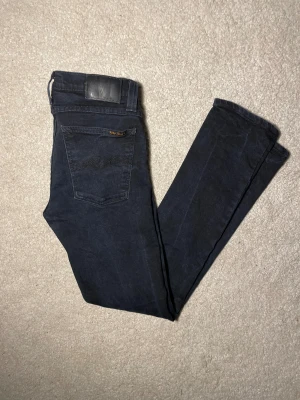 Svarta Nudie Jeans  W27L30 - Säljer ett par svarta jeans från Nudie Jeans i storlek W27 L30. De har klassisk femficksdesign, svarta sömmar och Nudie-logga på bakfickan. Jeansen är tillverkade i stretchig bomull för en bekväm och tight passform.