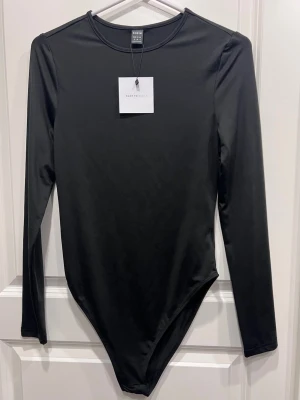 Svart långärmad body från SHEIN - Minimalistisk svart body från SHEIN i storlek M. Modellen har rund halsringning och långa ärmar, perfekt för att skapa en clean och stilren look. Materialet är mjukt och stretchigt syntet som sitter skönt mot kroppen. Enkel att matcha med jeans eller kjol.