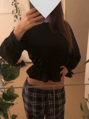 Svart blus med volang och knyt - Säljer en svart blus med volangkant nedtill och knyt vid ärmsluten. Blusen har långärmade puffiga ärmar och en croppad passform som ger en snygg siluett. Perfekt att styla med höga jeans eller kjol.