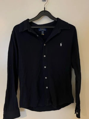 Svart långärmad skjorta från Polo Ralph Lauren - Svart skjorta från Polo Ralph Lauren med klassisk krage och vita knappar framtill. Liten broderad logga i vitt på bröstet. Skjortan är långärmad och har knappslå vid ärmslut. Tillverkad i mjuk bomull som känns skön mot huden.