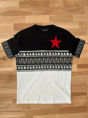 Givenchy svart och vit t-shirt med röd stjärna - Snygg t-shirt från Givenchy med svart överdel och vit nederdel. Framsidan har en stor röd stjärna på bröstet och coolt geometriskt mönster i grått och vitt över magen och ärmarna. Rund hals och korta ärmar. Perfekt för dig som gillar streetstyle och designer.