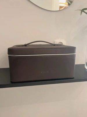 Brun sminkväska från CAIA - Säljer en stilren och rymlig brun sminkväska från CAIA med praktiskt handtag och silverfärgad dragkedja. Väskan har en rektangulär form och insidan har elastiska fack för penslar eller små tillbehör. Perfekt för att organisera dina skönhetsprodukter.