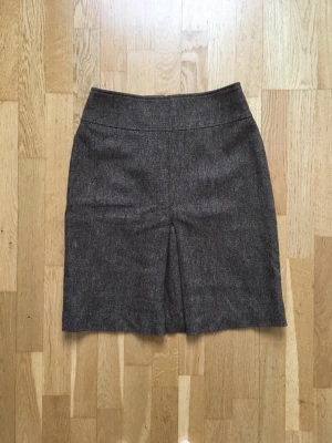 Vintage ullkjol - Fin vintage kjol i ullblandning från Hennes. Kjolen har ett stiligt veck fram och stängs med dragkedja bak. Tyget är en brunmelerad ullblandning med inslag av olika färger. Kjolen har ett foder som hindrar yttertyget från att stickas eller fastna på strumpbyxorna. Längd: 55cm. 