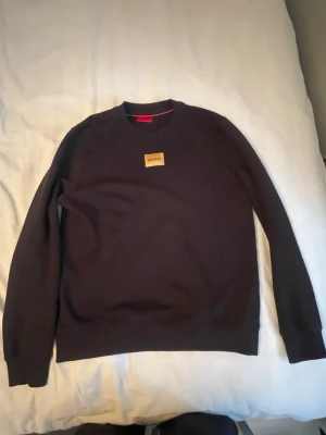 Svart sweatshirt från Hugo boss - Säljer en svart sweatshirt från Hugo boss med rund hals och en snygg beige patch med loggan på bröstet. Tröjan har långa ärmar och en klassisk passform. Perfekt till chill vardagsstil.