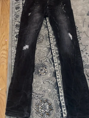 Svarta slim fit jeans från Dsquared2 - Svarta jeans från Dsquared2 med slim passform och slitna detaljer. Jeansen har en cool tvätt med ljusare partier och små färgstänk, samt trasiga partier på benen. Materialet är klassisk jeans och knapparna har Dsquared2-logga.