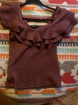 Plommonlila topp med volang från Lindex - Säljer en plommonlila topp från Lindex i storlek M. Toppen har en offshoulder-design med bred volang runt axlarna och ett krinklat tyg. Använd sparsamt. 