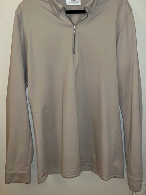 Beige långärmad half zip från Ciszere - Snygg beige långärmad tröja från Ciszere i storlek L, slim fit. Tröjan har en krage och en half zip-dragkedja framtill. Tillverkad i mjuk bomull som känns skön mot huden. Perfekt för dig som gillar stilrena och enkla plagg. Sitter mer som en M