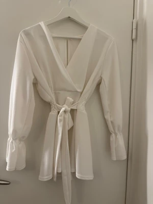 Vit omlottklänning med knytband - Supersnygg vit kortklänning med omlott-design och v-ringad hals. Klänningen har långa ärmar med lätt puff och resår vid ärmslut, samt ett knytband i midjan som ger en fin siluett. Materialet känns mjukt och har ett lätt fall. Perfekt för dig som gillar stilrena och trendiga plagg.