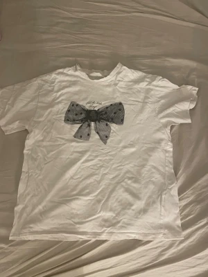 Vit t-shirt med svart rosett från H&M - Vit t-shirt från H&M i bomull med korta ärmar och rund hals. På bröstet sitter en stor svart rosett i mesh med prickigt mönster, vilket ger en unik och cool look. Perfekt för dig som gillar statement-detaljer och vill sticka ut lite extra.