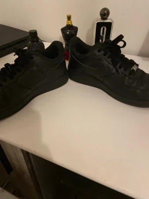 Nike Air Force 1 helsvarta sneakers - Säljer ett par klassiska Nike Air Force 1 sneakers i helsvart färg. Skorna har rund tå, snörning och är tillverkade i skinn med platt sula. Perfekta för dig som gillar stilrena och tidlösa sneakers med ikonisk design. Få antal gånger använd och är i ganska bra skick. Storlek 42 för dig som gillar sådana svarta Nike Airforces. 