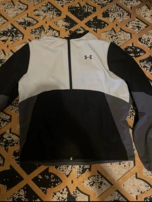 Svart och vit vindjacka från Under Armour - Säljer en snygg vindjacka från Under Armour i svart, vitt och grått. Jackan har huva med snörning, hel dragkedja framtill och meshfoder för extra komfort. Perfekt för dig som vill ha en sportig look och gillar funktionella detaljer.
