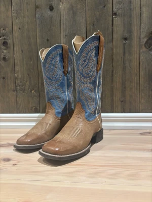 Blå och bruna cowboy boots från Ariat - Säljer ett par snygga cowboy boots från Ariat (Shock Sheild) med blå skaft och bruna detaljer. Bootsens skaft har ett paisley-inspirerat broderat mönster och är tillverkade i skinn och textil. De har en fyrkantig tå och robust klack, perfekta för dig som gillar westernstil. Endast använda 4 gånger men är för små för mig! 2200kr nya och kommer med orginalboxen