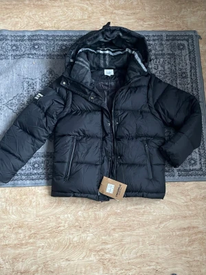 Svart pufferjacka från Burberry - Säljer en svart pufferjacka från Burberry med huva och stora fickor framtill. Jackan har Burberry London England-patch på ärmen och dragkedja framtill. Insidan har ett rutigt foder i huvan och jackan är riktigt fluffig och varm. Perfekt för kalla dagar.
