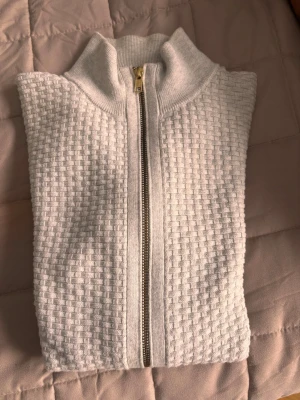 Vit stickad kofta från John Henric - Säljer en vit stickad kofta från John Henric med snyggt strukturerat rutmönster och guldfärgad dragkedja framtill. Koftan har en hög krage och är perfekt för lager-på-lager under kyliga dagar. Passar dig som gillar stilrena och klassiska plagg.