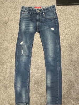 Blå skinny jeans med slitningar - Säljer ett par blå skinny jeans från Supersoft med coola slitningar och detaljer på framsidan. Jeansen har klassisk femficksdesign, dragkedja och knapp, samt en snygg tvättad look. Perfekt för dig som gillar en smal passform och streetstil.