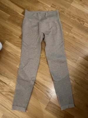 Beige seamless leggings från Aim'n - Säljer ett par beige seamless leggings från Aim'n med ribbad hög midja och snyggt strukturerat mönster på benen. Materialet är mjukt och stretchigt, perfekt för träning eller chill. Metallmärke med Aim'n-logga vid midjan. Tight passform och diskret design. En maska vid ena benet (se bild).