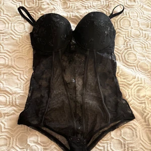 Svart spetskorsett/body från Intimissimi - Säljer en svart OANVÄND med prislapp korsett/body från Intimissimi med vadderade kupor och justerbara axelband. Toppen är i transparent spets med blommönster och har dekorativa sömmar framtill. Knäppning i grenen och snyggt glittriga detaljer för extra effekt.