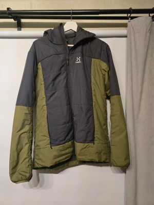 Grön och mörkgrå pufferjacka Haglöfs - Säljer en pufferjacka från Haglöfs i mörkgrått och olivgrönt. Jackan har huva, lång ärm och dragkedja framtill. Perfekt för kyliga dagar och outdoor-aktiviteter. Materialet är syntet och polyester, vilket gör den lätt och varm. Snygg och funktionell design.