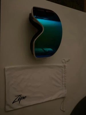 Ingen sko på bilderna - Bilderna visar ett par skidglasögon från Zipe med vit förvaringspåse, inte skor. Glasögonen har blåtonad lins och vit ram med bredt vitt band.