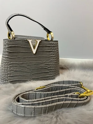 Grå handväska med krokodilmönster - Snygg grå handväska med krokodilmönstrad yta och guldfärgade detaljer. Väskan har ett stort V i metall framtill och kommer med både handtag och avtagbar axelrem. Perfekt för dig som gillar stilrena accessoarer med lite edge.