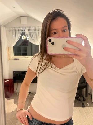 Vit croppad topp med draperad hals - Säljer en vit croppad topp med korta ärmar och snygg draperad halsringning, som är köpt i Thailand. Toppen har en tight passform och är tillverkad i ett mjukt bomullsmaterial. Perfekt till jeans eller kjol för en fräsch look. Obs står ej någon storlek, men passar som en s-xs