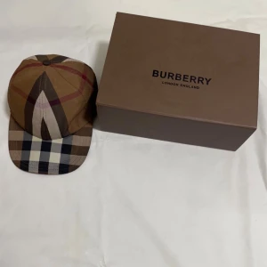 Rutig Burberry keps i bomull - Säljer en snygg Burberry keps i klassiskt rutigt mönster med bruna, beige, svarta och vita toner. Kepsen är tillverkad i 100% bomull och har justerbar rem med metallspänne där Burberry är ingraverat. Perfekt för dig som vill ha en ikonisk accessoar.