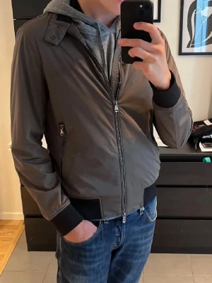 Brun bomberjacka med dragkedja - Snygg grå bomberjacka med svarta muddar och detaljer. Jackan har dragkedja framtill, två sidofickor med dragkedja och en cool knappdetalj vid axeln. Materialet är slätt och känns lätt, perfekt för dig som gillar stilren och enkel look. 