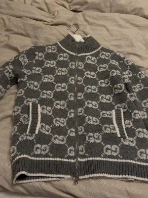 Grå kofta med dragkedja från Gucci - Säljer en grå stickad kofta från Gucci med vit GG-mönster över hela plagget. Koftan har ribbad krage, muddar och nederkant samt två fickor med vit kant. Dragkedja framtill och mjukt material som håller dig varm.