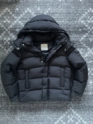Svart Moncler pufferjacka - Svart pufferjacka från Moncler med stor huva, quiltad design och klassisk Moncler-logga på ärmen. Jackan har dragkedja och tryckknappar framtill samt praktiska fickor. Insidan har en unik serietidningsetikett. Perfekt för kalla dagar.