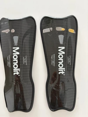 Monolit benskydd i kolfiber - Säljer ett par svarta benskydd 19cm från Monolit, tillverkade i kolfiber med ergonomisk form och vadderad insida för extra komfort. Perfekta för dig som spelar fotboll och vill ha lättviktsskydd med snygg design. Passar dig som gillar sportig och modern utrustning.