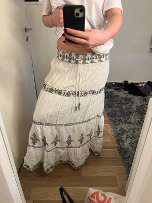 Zara långkjol med broderade detaljer - Säljer en vit långkjol med svarta broderade mönster och snörning i midjan. Kjolen har flera dekorativa band med blommiga broderier och är tillverkad i ett lätt, skrynkligt bomullstyg. Perfekt för dig som gillar boho-stil och vill ha något luftigt och snyggt.