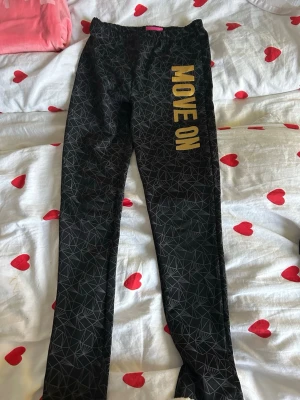 Svarta leggings med gult tryck från Stage - Säljer ett par svarta leggings från Stage med geometriskt mönster i grått och stort gult tryck 'MOVE ON' längs benet. De har elastisk midja och är gjorda i mjukt syntetmaterial, perfekta för träning eller chill. Supersnygga och bekväma!