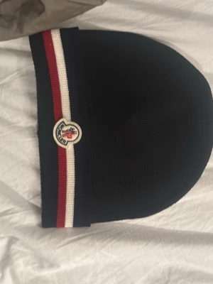 Svart Moncler mössa med röd och vit rand - Snygg svart Moncler mössa med klassiska ränder i rött och vitt längs kanten och Moncler-logga framtill. Mössan är ribbstickad och har en stilren, enkel form som passar perfekt till vintern. Materialet är mjukt och värmande. Pris kan diskuteras!!!