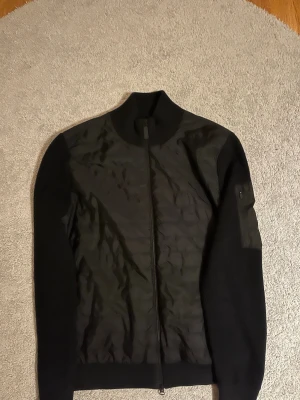 Svart bomberjacka från J.Lindeberg - Svart bomberjacka från J.Lindeberg med ribbad krage och ärmar. Jackan har quiltad front i syntetmaterial och en ficka med dragkedja på ärmen. Snygg och stilren design som passar perfekt till streetwear och vardagsstil.