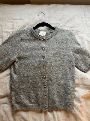 Grå kofta med guldfärgade knappar H&M - Säljer en grå stickad kofta från H&M i storlek XS. Koftan har korta ärmar, rund halsringning och snygga guldfärgade knappar framtill. Perfekt att slänga över en topp för en chill men ändå stilsäker look.