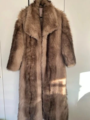 Lång brun pälsjacka med bred krage - Säljer en riktigt fluffig och lång (till anklarna om du är 172 eller kortare) pälsjacka i brun/beige nyans med bred krage. Jackan har en lyxig och maffig look, perfekt för dig som vill sticka ut och hålla dig varm. Den har en rak passform och täcker hela kroppen, vilket gör den extra mysig under kalla dagar.