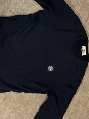 Svart tröja från Stone Island - Svart långärmad sweatshirt från Stone Island med klassisk rund hals och ikonisk logopatch på bröstet. Tröjan har en clean look och är gjord i mjuk bomull som känns skön mot huden. Perfekt för dig som gillar streetwear och vill ha något stilrent.
