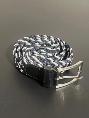 Flätat bälte 105 cm - Flätat bälte som är 105 cm långt, men materialet är stretchigt. Bältet är i färgerna vitt, svart och blått. Aldrig blivit använt. 