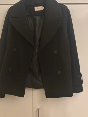 Svart skepparkavaj från Nelly XXS - Stilren svart skepparkavaj från Nelly i storlek XXS. Jackan har dubbelknäppning framtill, bred krage och snygga detaljer med knappar på ärmsluten. Perfekt för dig som gillar klassisk och cool look. Pris kan diskuteras!!