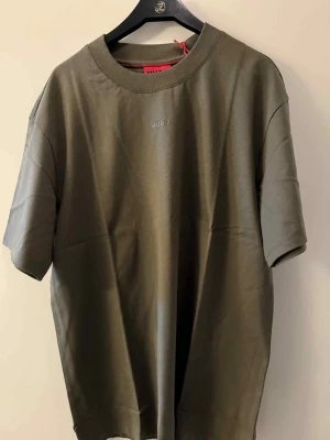 Olivgrön t-shirt från HUGO, M - Snygg olivgrön t-shirt från HUGO med diskret logga på bröstet. Modellen har rund hals och korta ärmar, tillverkad i mjuk bomull som känns skön mot huden. Passformen är loose och t-shirten har en clean look utan mönster eller extra detaljer.