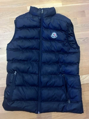 Moncler väst - Säljer en svart dunväst från Moncler med klassisk logga på bröstet. Västen har quiltad design, två fickor med dragkedja och hög krage. Perfekt att slänga över en hoodie eller långärmad tröja när det är kyligt ute. Snygg och stilren look.