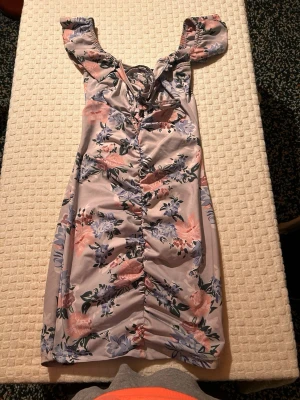 Blommig kortklänning med snörning - Säljer en söt kortklänning med blommigt mönster i ljusa toner av rosa, lila och grönt. Klänningen har snörning framtill, volangdetaljer vid axlarna och är tillverkad i ett mjukt syntetmaterial. Perfekt för sommarens alla tillfällen!