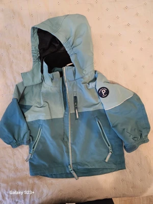 Blå vindjacka Polarn O. Pyret 98 - Snygg blå vindjacka från Polarn O. Pyret med huva och flera dragkedjor. Jackan har elastiska muddar, reflexdetaljer och meshfoder på insidan. Perfekt för blåsiga dagar och har praktiska fickor med dragkedja. Märke på ärmen och justerbar nederkant.