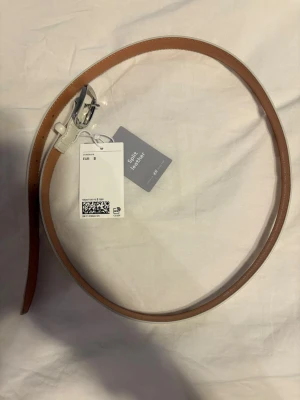 Vit och beige skinnbälte från H&M - Säljer ett stilren bälte från H&M i split leather med vit framsida och beige insida. Bältet har ett klassiskt silverfärgat spänne och är enkel att matcha med olika outfits. Perfekt för dig som gillar minimalistisk och clean stil. Pris är diskuterbart!
