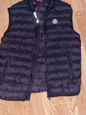 Svart dunväst från Moncler - Säljer en svart dunväst från Moncler med quiltad design och dragkedja framtill. Västen har två sidofickor med dragkedja och Moncler-logga på bröstet. Insidan har coola etiketter och ett serietryck. Perfekt för dig som vill ha en snygg och varm väst. Storlek S