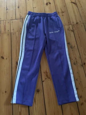 Äkta Palm Angels Byxor - Säljer ett par lila mjukisbyxor från Palm Angels med vita sidoremsor och svarta detaljer. Byxorna har elastisk midja, raka ben och dragkedjor vid bensluten. Palm Angels-loggan är broderad på framsidan. Perfekt för en chill och sportig look.