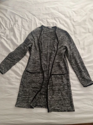 Gråmelerad lång kofta från me&i - Lång och loose gråmelerad kofta från me&i i storlek M. Koftan har svarta kantdetaljer, två fickor framtill och är långärmad. Perfekt att slänga på sig när det är lite kyligt ute. Materialet känns mjukt och stickat.