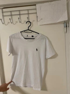 Vit t-shirt från Ralph Lauren - Vit t-shirt från Ralph Lauren med klassisk rund hals och korta ärmar. På bröstet finns den ikoniska Polo-loggan i svart. T-shirten är gjord i mjuk bomull och har en clean, stilren look som funkar till allt. Perfekt för dig som gillar enkel och snygg stil. Liknande modell som original. 
