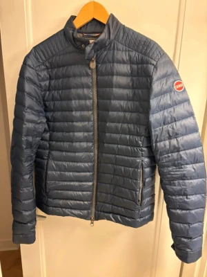 Mörkblå pufferjacka från Colmar - Snygg mörkblå pufferjacka från Colmar med quiltad design och hög krage. Jackan har en tydlig logga på ärmen, dragkedja framtill och två sidofickor med dragkedja. Perfekt för dig som vill ha en stilren och lätt jacka med sportig vibe.